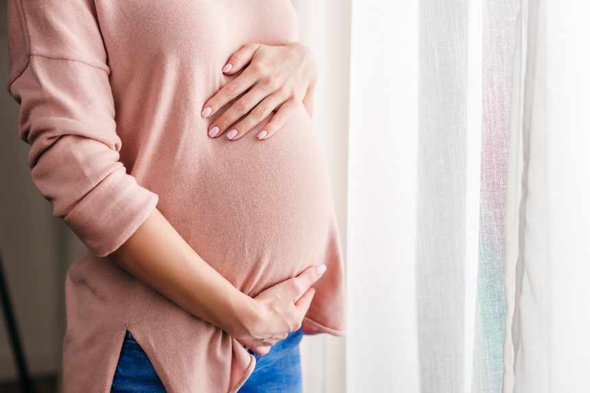 pregnant woman standing by sunny window and gently holding her tummy